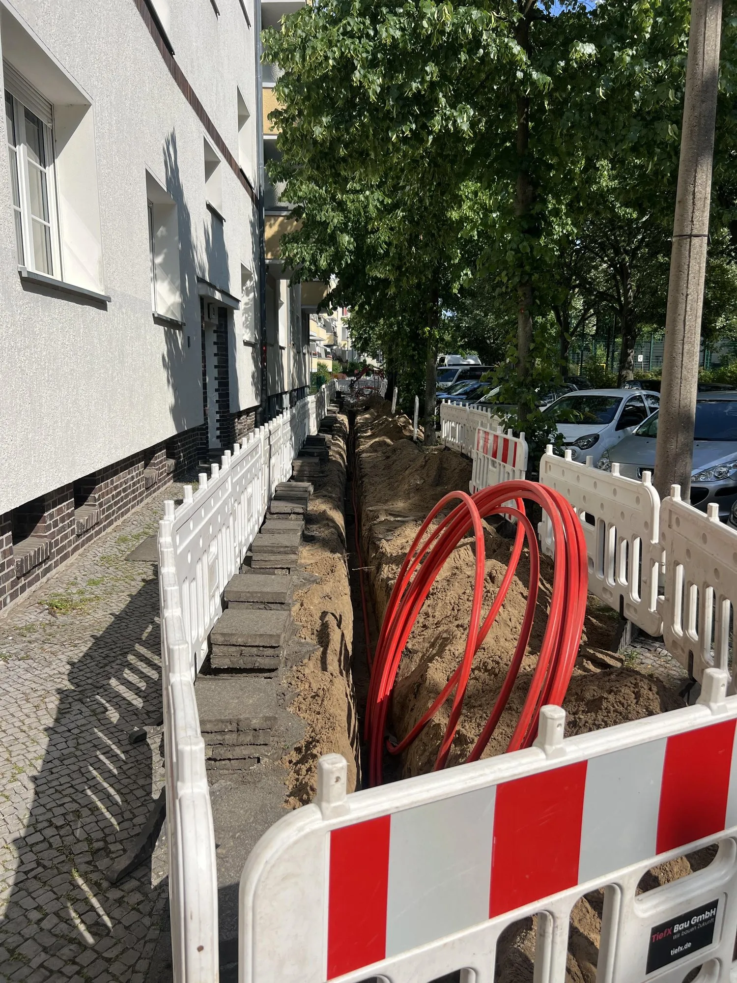 Offener Leitungsgraben im Gehweg eines Berliner Wohngebiets, abgesichert mit rot-weißen Absperrgittern der TiefX Bau GmbH, daneben aufgerollte rote Schutzrohre
